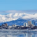 The image shows the winter skyline of downtown Anchorage, Alaska, set against the snowy Chugach Mountains and the icy Cook Inlet.