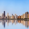The image shows the Chicago skyline with the John Hancock Center visible on the left, reflected in the calm waters of Lake Michigan along the lakefront.
