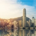 The image shows the iconic skyline of Victoria Harbour in Hong Kong during golden hour, reflecting brilliantly on the calm water's surface.