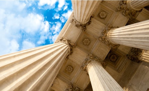 The image displays a low-angle view of the neoclassical marble columns at the Supreme Court of the United States in Washington, D.C.
