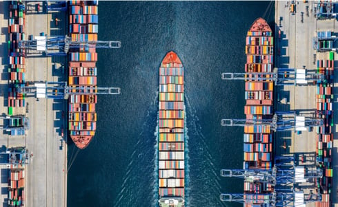 An aerial view shows a large container ship moving through a narrow channel between busy port terminals lined with stacked shipping containers and gantry cranes.
