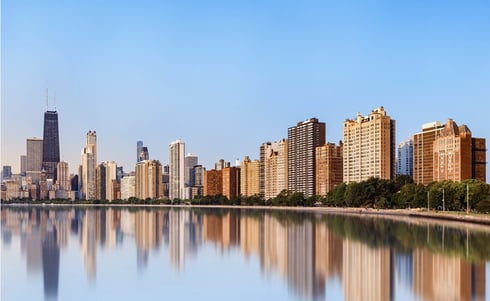 The image shows the Chicago skyline with the John Hancock Center visible on the left, reflected in the calm waters of Lake Michigan along the lakefront.